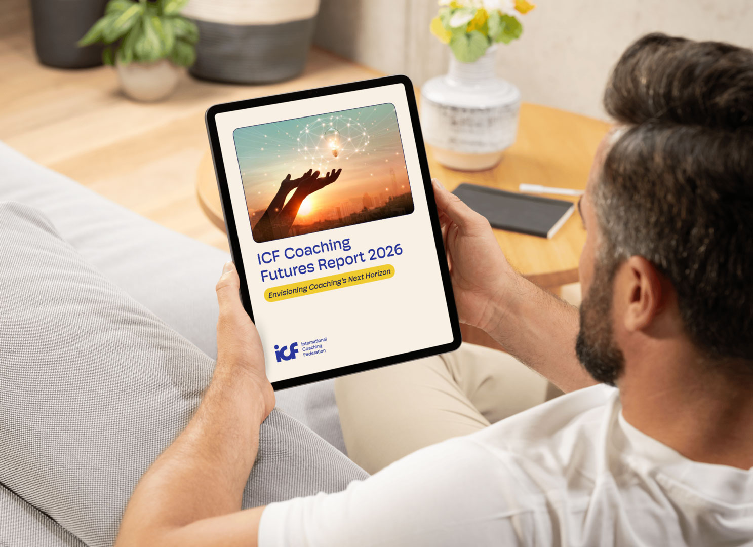 A man with dark hair and a beard wearing a white tshirt sits on a sofa holding a tablet